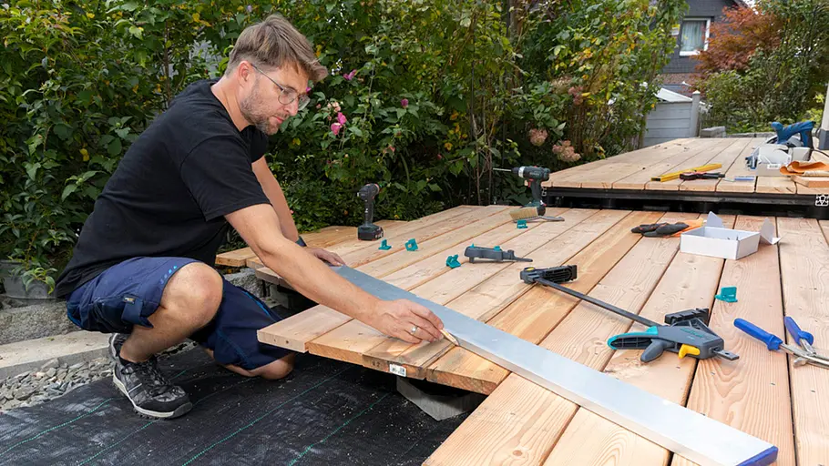 Terrasse gestalten Ein Mann nutzt eine lange Aluschiene als Führung, um den Versatz der Terrassendielen für den Endzuschnitt anzureißen.