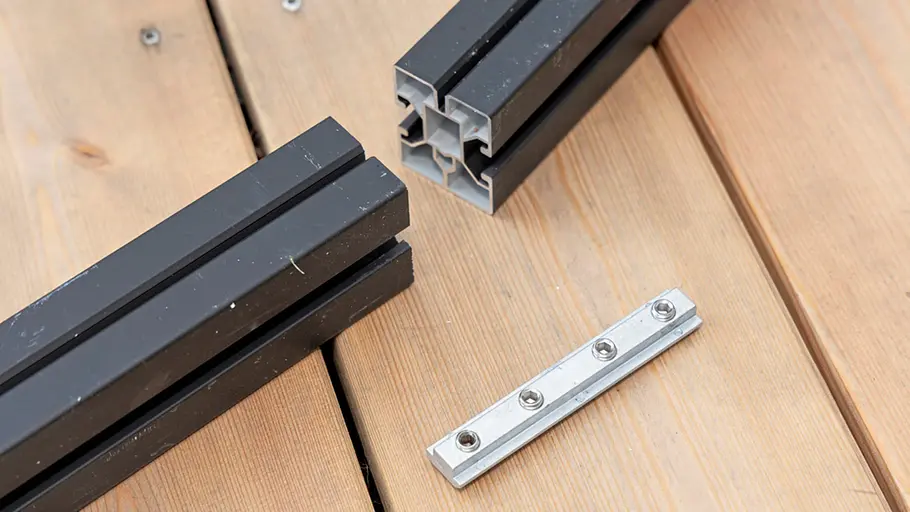 Terrasse gestalten Zwei schwarze Aluminiumprofile für die Terrassenunterkonstruktion und eine silberne Verbindungsschiene liegen auf Holzdielen.
