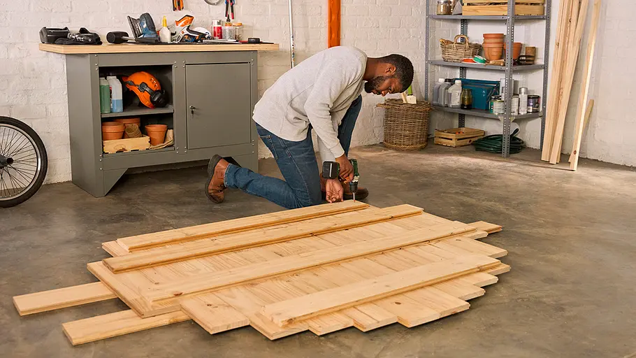 Ein Mann kniet in einer Werkstatt und schraubt mehrere Holzbretter zu einer größeren Holzkonstruktion zusammen.