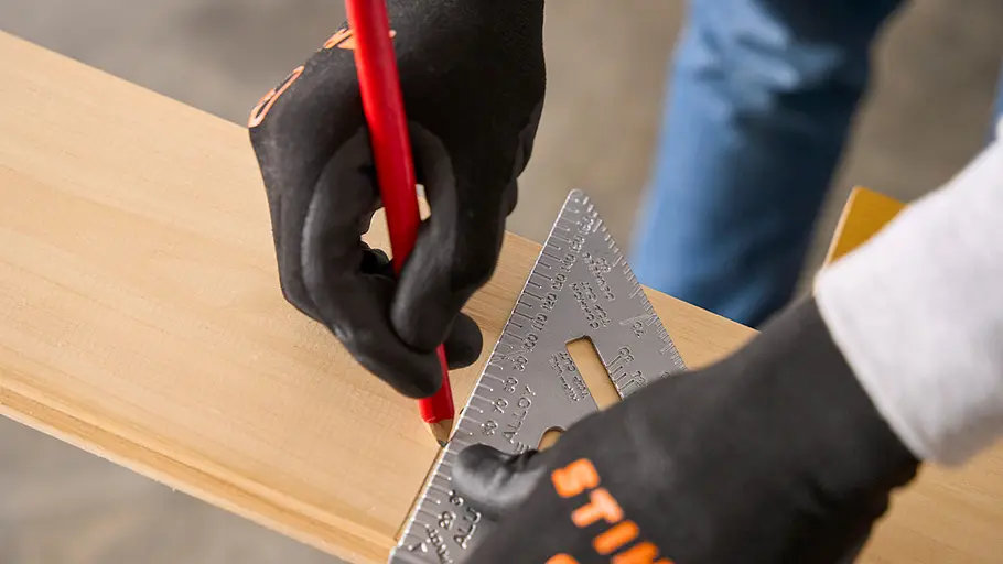Eine Person mit schwarzen Handschuhen zeichnet mit Bleistift und Winkelmaß eine Linie auf ein Holzbrett.