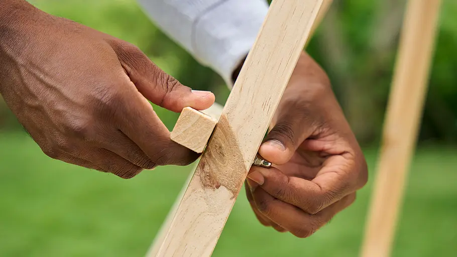 Zwei Hände befestigen mit einer Flügelschraube und einem Holzklotz eine Querlatte an einer Tipi-Stange im Garten.