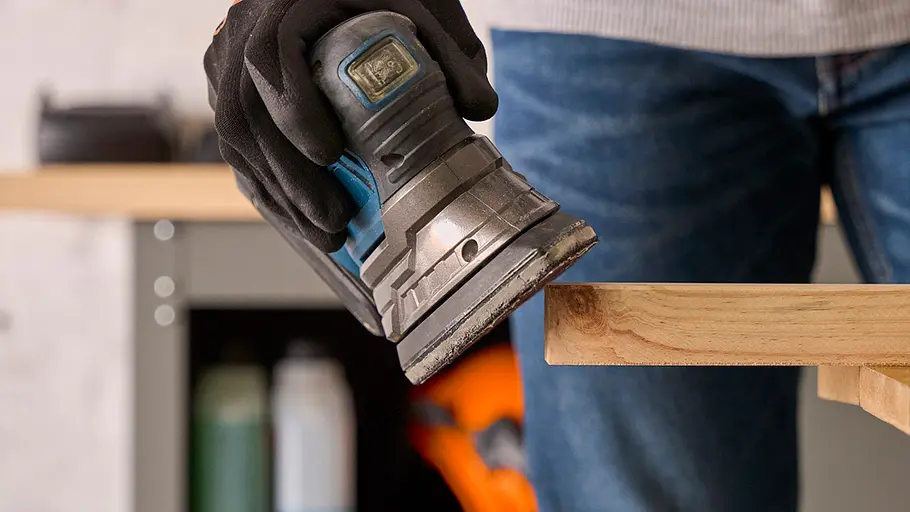 Eine Person hält einen elektrischen Schleifer und bearbeitet die Kante eines hellen Holzbretts in einer Werkstatt.