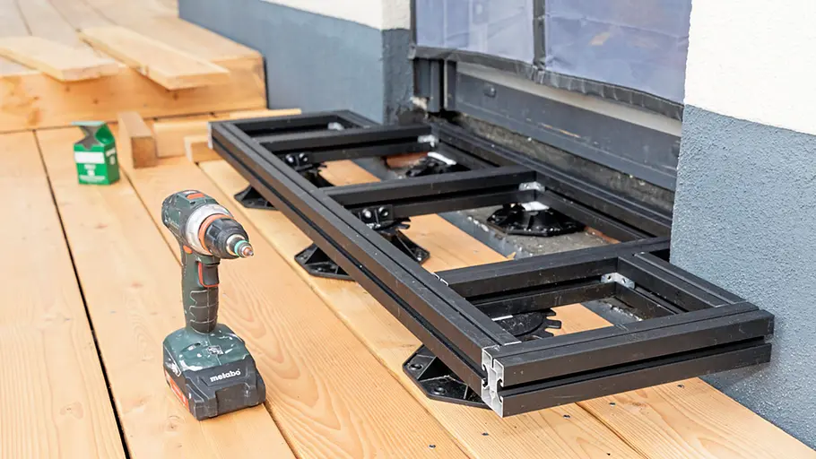 Terrasse gestalten Eine Bohrmaschine steht auf Holzplanken, daneben liegt eine schwarze Metallunterkonstruktion vor einer Terrassentür.
