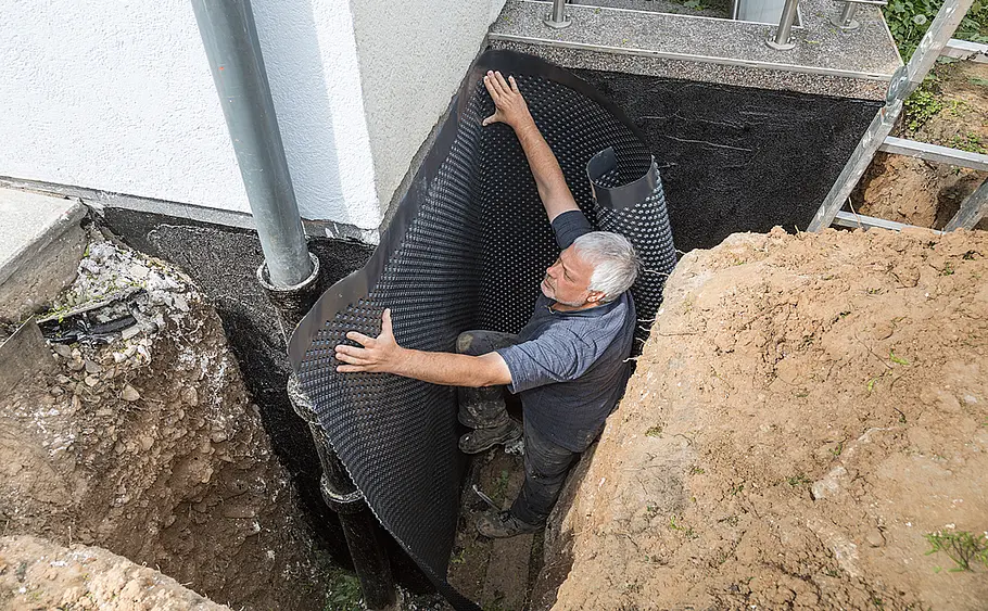 Anbringen von Noppenfolie an abgedichteter Hauswand Ein Mann bringt in einer Baugrube Noppenfolie an einer neu abgedichteten Hauswand an.