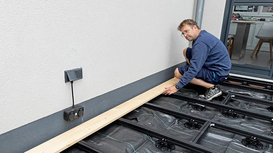 Terrasse gestalten Ein Mann legt ein Holzbrett auf die vorbereitete Unterkonstruktion der Terrasse an einer Hauswand an.