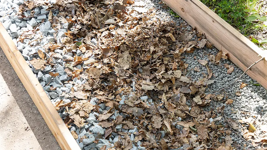 Ein Holzkasten ist mit einer Schicht aus Laub und grobem Kies gefüllt und wird von Sonnenlicht beleuchtet.
