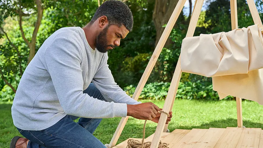 Ein Mann kniet auf dem Rasen und befestigt eine dicke Kordel an einer Tipi-Stange, das Zelttuch liegt gerafft am Gestell.