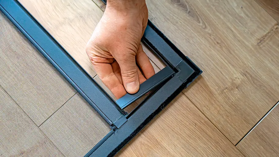 Eine Hand setzt am unteren Rahmen einer Insektenschutztür ein rechteckiges schwarzes Kunststoffteil auf hellen Holzboden ein.