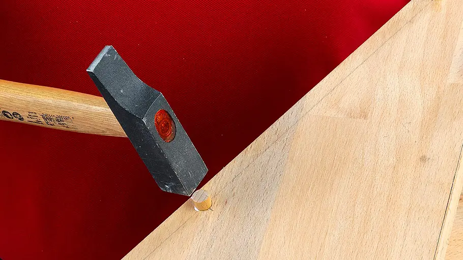 Verdrehter Hocker Ein Hammer treibt einen Holzdübel in ein gebohrtes Loch einer dreieckigen Holzplatte vor rotem Hintergrund.