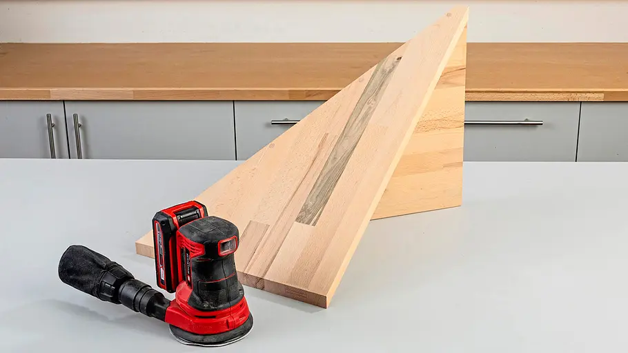 Verdrehter Hocker Ein zusammengeschraubtes, dreieckiges Holzelement steht auf der Werkbank, davor liegt ein roter Akku-Exzenterschleifer.