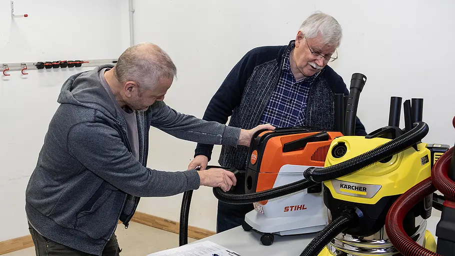 Werkstattsauger Begutachtung Zwei Männer testen einen orangefarbenen und einen gelben Werkstattsauger an einem Tisch in heller Werkstattumgebung.