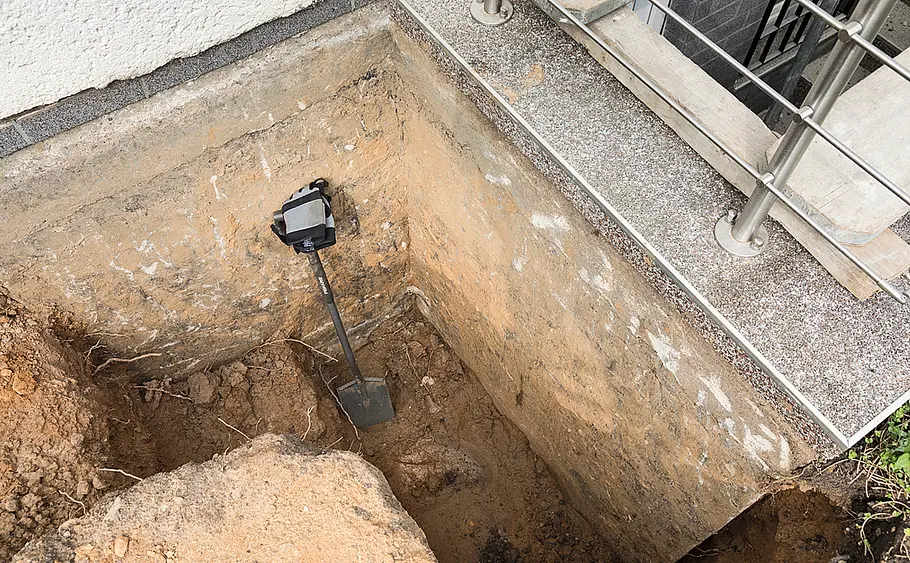 Ausgehobene Grube an Hauswand mit Spaten Eine ausgehobene Baugrube an der Hauswand, ein Spaten steht aufrecht im Erdreich am Rand der Grube.