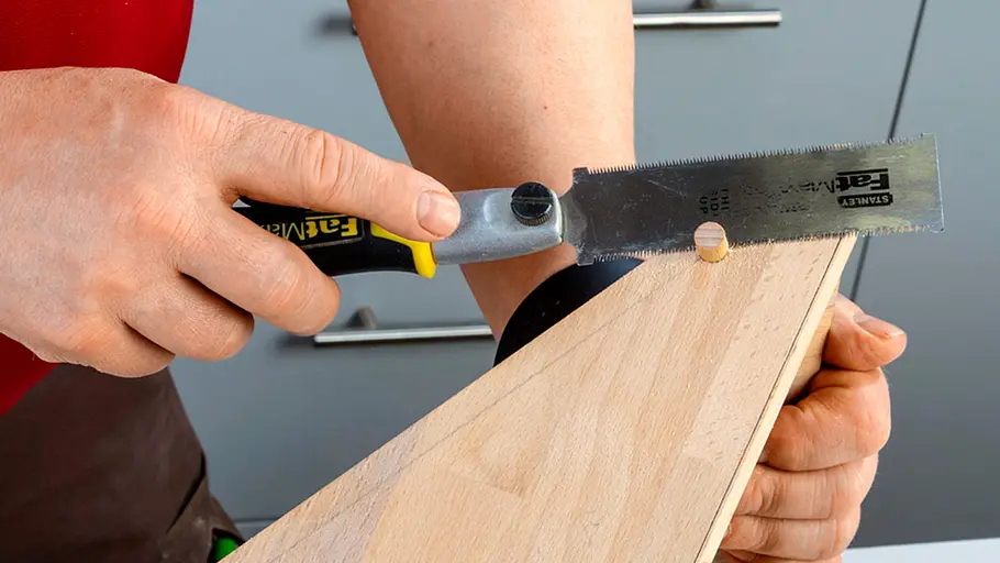 Verdrehter Hocker Eine Person sägt mit einer Feinsäge den überstehenden Teil eines Holzdübels an einer Holzplatte ab.