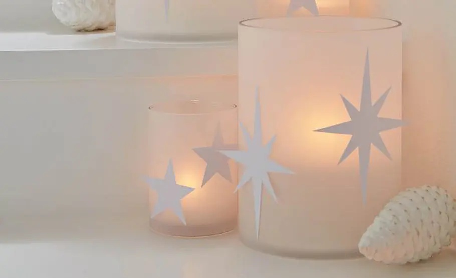 Weihnachts-Windlicht: Funkelndes Sternen-Glas Zwei Windlichter mit weißen Sternmotiven leuchten auf einer hellen Treppenstufe neben einer Deko-Zapfe.