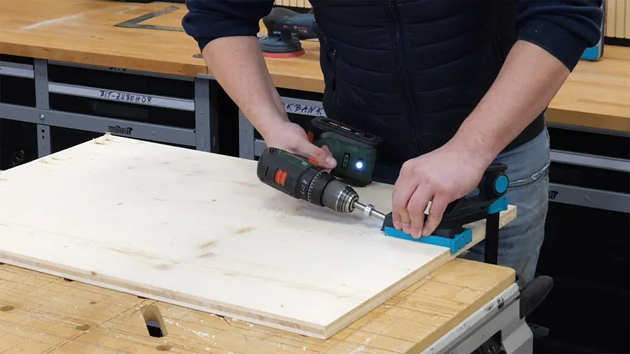 Paketbox Eine Person bohrt mit einer Bohrmaschine ein Loch in ein helles Holzbrett, das an der Kante fixiert ist.
