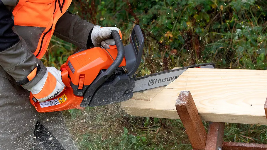 Praxistest für die Benzin-Kettensägen Eine Person mit orangefarbener Schutzkleidung sägt mit einer Husqvarna-Motorsäge ein helles Holzbrett im Freien.
