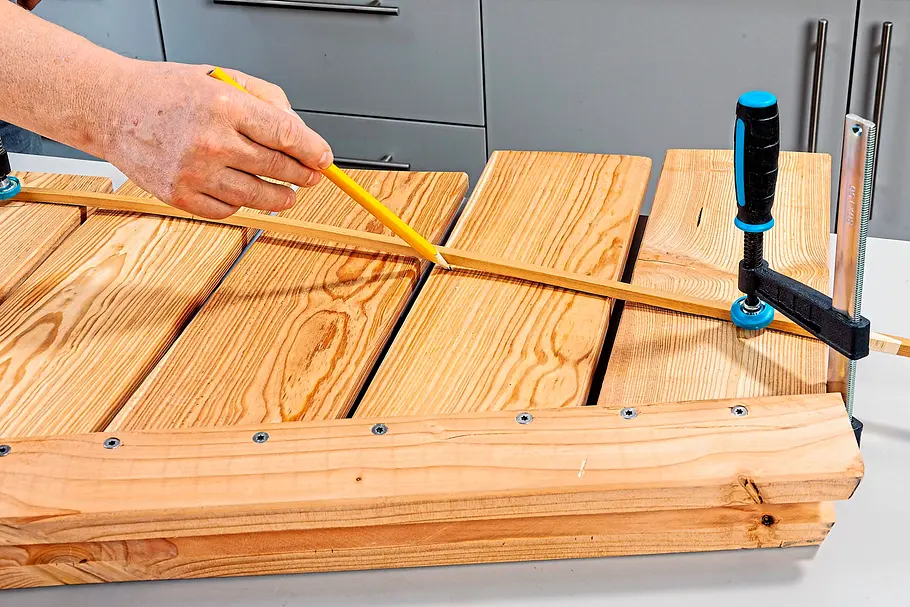 Gartendusche bauen Eine Person zeichnet den Verlauf eines Kupferrohrs auf einer aus Holzlatten zusammengesetzten Fläche an, die mit einer Schraubzwinge fixiert ist.
