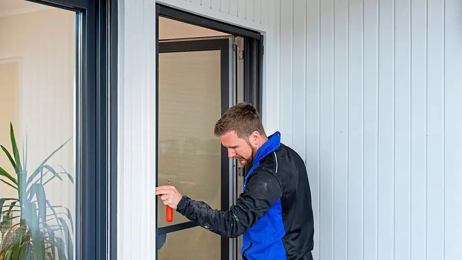 Ein Mann in blauer Arbeitsjacke montiert mit einem Schraubendreher eine Insektenschutztür an einem weißen Hauseingang.