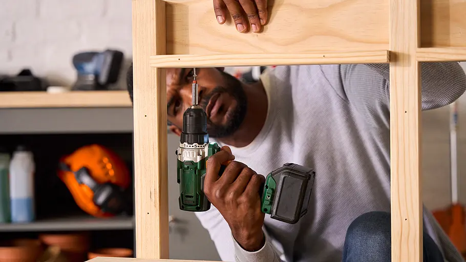 Matschküche: Regalbretter Ein Mann befestigt mit einer Akkubohrmaschine ein Regalbrett an der Rückwand der Matschküche aus Holz.