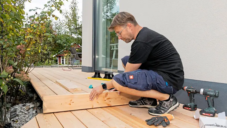 Terrasse gestalten Ein Mann setzt eine Holzblende als Abschluss an die Stirnseite der fertigen Holzterrasse an und richtet sie aus.