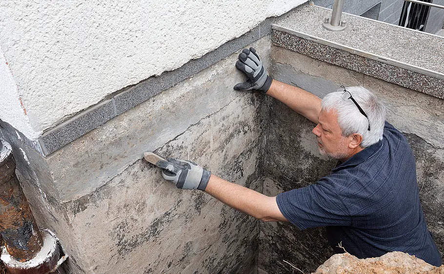 Vorbereitungsarbeiten an freigelegter Hauswand Ein Mann mit Handschuhen glättet mit einer Drahtbürste die freigelegte Hauswand in einer Baugrube.