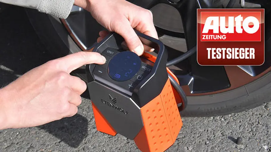 Zwei Hände bedienen eine orange-schwarze Akku-Luftpumpe am Reifen eines Autos; Auto Zeitung Testsieger-Logo sichtbar.