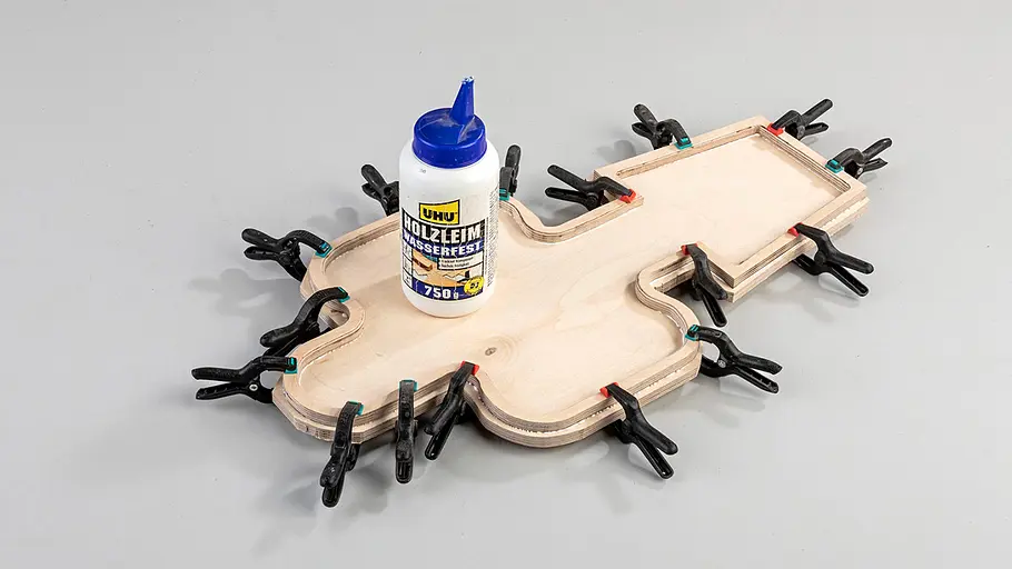 Brett und Rahmen leimen Mehrere Schraubzwingen fixieren zwei Kaktus-förmige Holzplatten, eine Flasche Holzleim steht darauf.