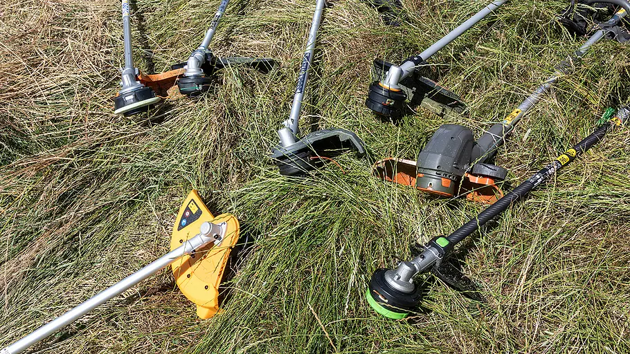 Mehrere Akku-Freischneider verschiedener Hersteller liegen auf einer Wiese mit hohem, geschnittenem Gras.