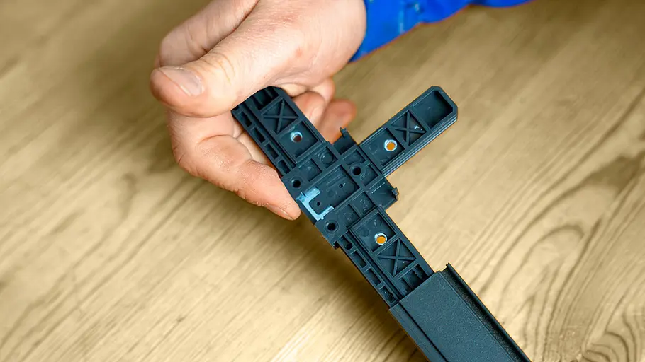 Eine Hand hält ein schwarzes Kunststoff-T-Stück, das an einem Rahmenprofil aus Aluminium befestigt ist.