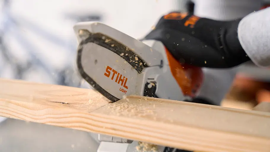 Matschküche: Latten zuschneiden Eine Person trägt Handschuhe und sägt mit einer STIHL-Kettensäge eine Holzlatte auf eine bestimmte Länge zu.