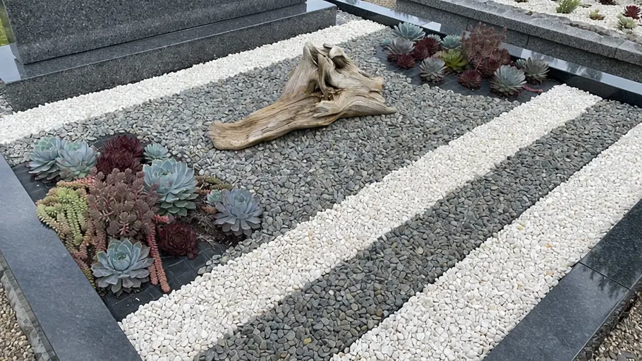Modernes Grab mit weißen und grauen Kiesstreifen, Sukkulenten und einem dekorativen Holzstück in der Mitte.
