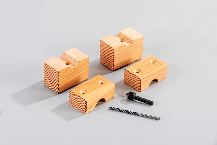 Gartendusche bauen Vier kleine Holzklötze mit eingefrästen Aussparungen sowie Schraube und Bohrer auf einer grauen Fläche.