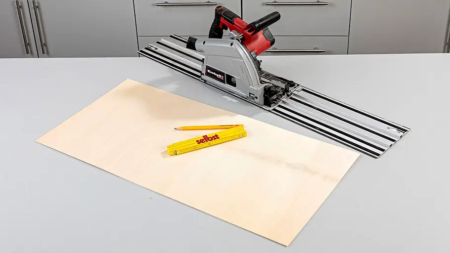 Platte zuschneiden Eine Tauchsäge mit Führungsschiene liegt hinter einer rechteckigen Holzplatte, davor Bleistift und Zollstock auf grauem Tisch.