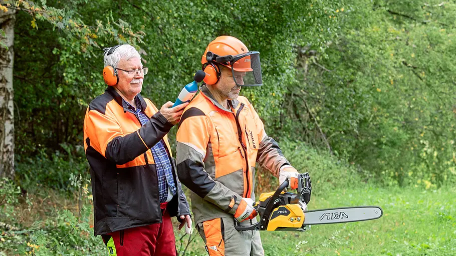Lautstärkemessung beim Motorsägen-Test Zwei Männer mit Gehörschutz stehen auf einer Wiese, einer hält ein Messgerät an den Gehörschutz des anderen mit Motorsäge.