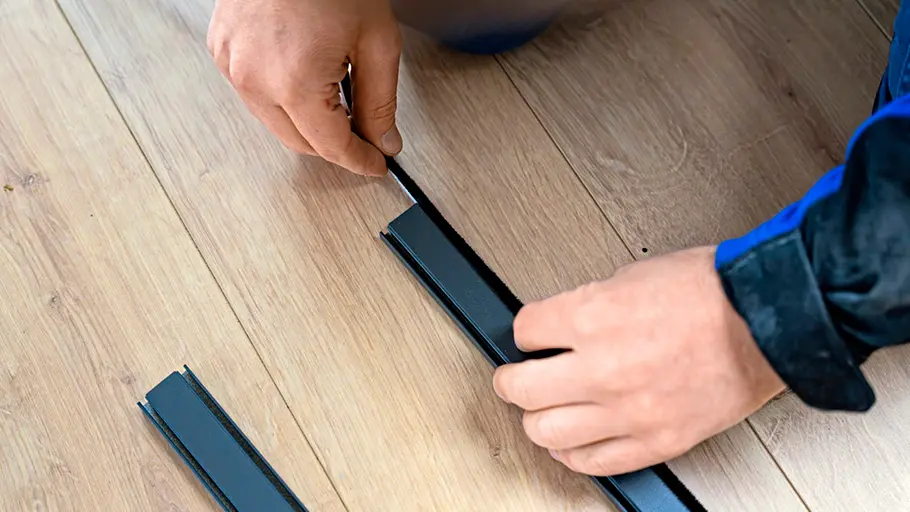 Zwei Hände setzen eine schwarze Bürstendichtung in einen Metallrahmen für eine Insektenschutztür auf einem Holzfußboden ein.