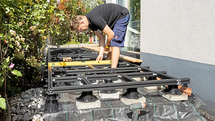 Terrasse gestalten Ein Mann richtet mit einer Wasserwaage das Metallgerüst der neuen Terrasse aus, umgeben von Pflanzen und Hauswand.