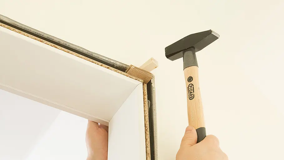 Türzarge ausrichten Eine Hand hält einen weißen Türrahmen, eine zweite Hand schlägt mit einem Hammer einen Keil am Türrahmen fest.