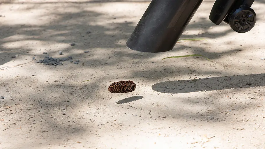 Die Düse eines Laubbläsers saugt einen Tannenzapfen auf hellem Sandboden an, im Hintergrund kleine Steinhaufen.