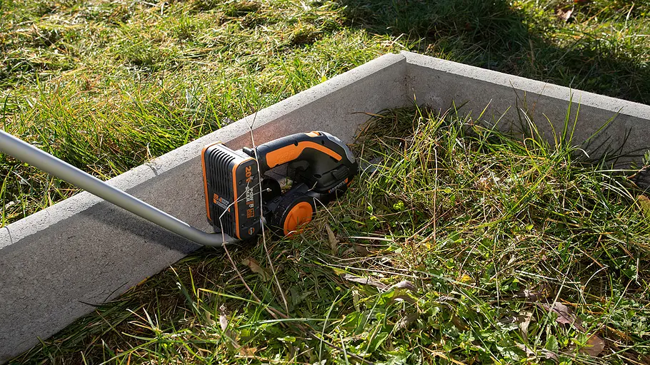 Akku-Grasscheren-Test: Rasenkante Eine schwarze und orangefarbene Akku-Grasschere schneidet entlang einer Rasenkante aus grauem Beton im Garten.