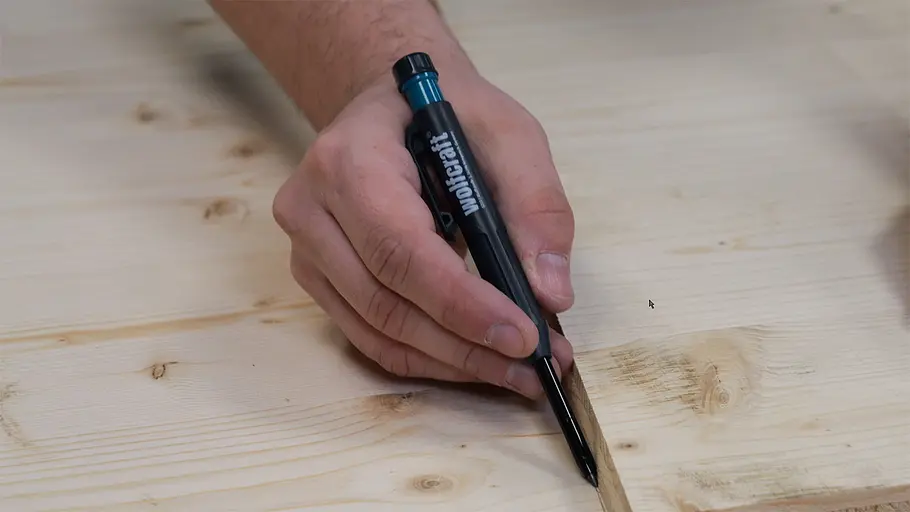Paketbox Eine Hand hält einen Stift und markiert eine gerade Linie entlang der Kante eines hellen Holzbretts.