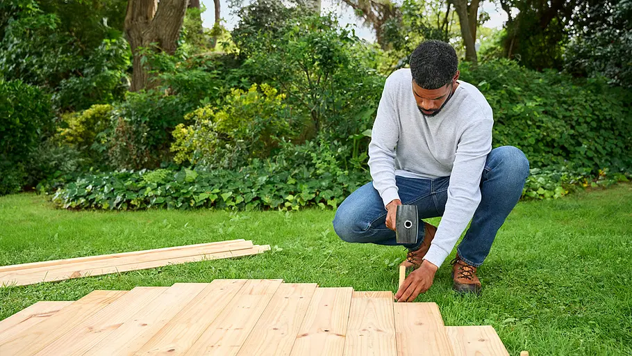 Ein Mann kniet auf einer Wiese und baut mit Holzlatten und einem Gummihammer eine Holzkonstruktion im Garten zusammen.