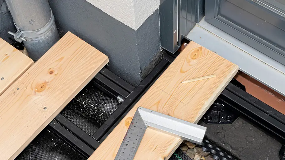 Terrasse gestalten Ein Bleistift und ein Winkel liegen auf einer markierten Holzdiele, die an eine Hauswand und ein Fenster angepasst werden soll.