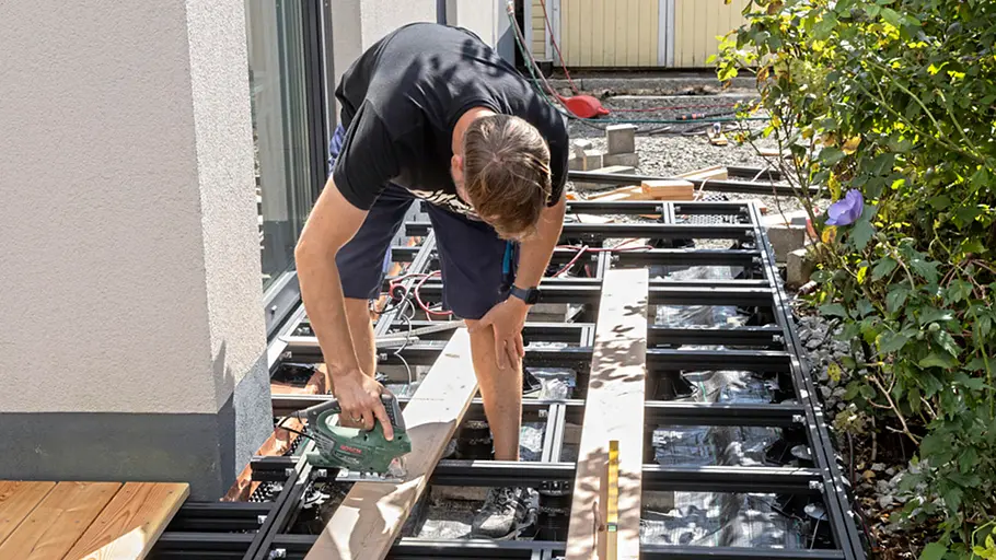 Terrasse gestalten Ein Mann sägt eine Terrassendiele auf der Unterkonstruktion passgenau mit einer Stichsäge direkt am Haus.