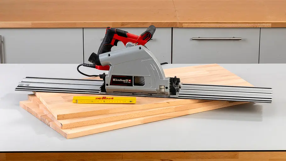 Verdrehter Hocker Eine Handkreissäge mit Führungsschiene, drei Holzplatten und ein Zollstock liegen nebeneinander auf einer Werkbank.