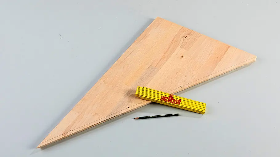Verdrehter Hocker Ein zugeschnittenes Holzbrett in Dreiecksform liegt auf einer Fläche, daneben liegen ein Zollstock und ein Bleistift.