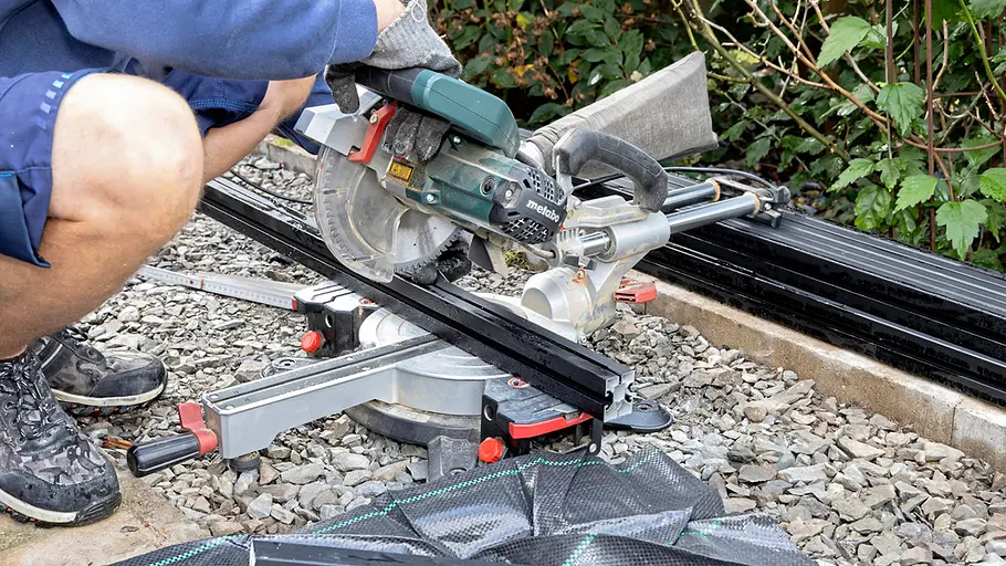 Terrasse gestalten Eine Person sägt mit einer Kappsäge ein schwarzes Metallprofil auf einer Kiesfläche im Garten passend zurecht.