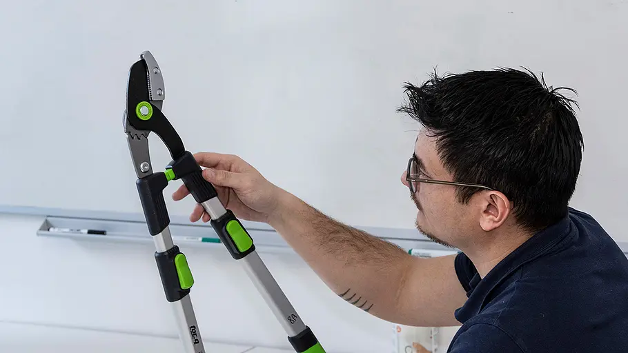 Astscheren-Test Begutachtung Ein Mann mit Brille prüft eine Astschere mit grün-schwarzen Griffen vor einer weißen Tafel im Raum.
