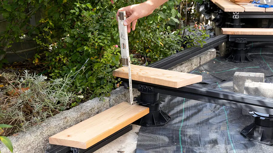 Terrasse gestalten Eine Person misst mit einem Zollstock den Abstand zwischen zwei Holzstufen auf der Terrassenkonstruktion ab.