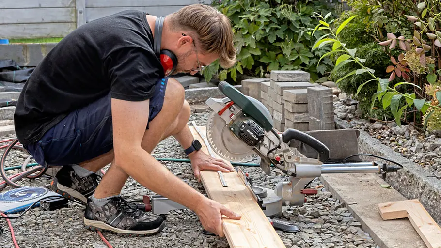 Terrasse gestalten Ein Mann sägt mit einer Kappsäge ein Holzbrett auf Maß, um die Terrasse passend zu gestalten.