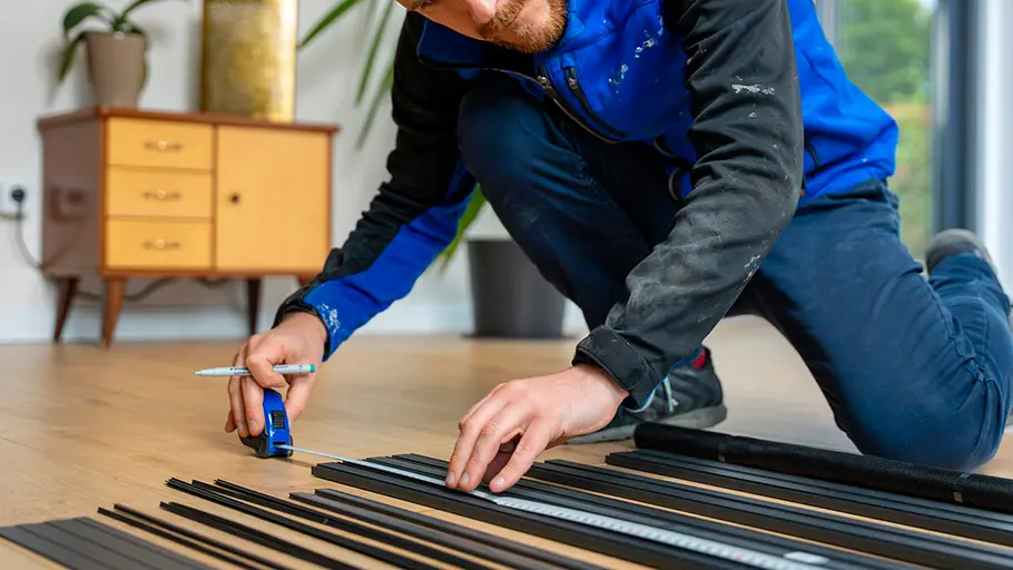 Ein Mann in blauer Arbeitsjacke misst schwarze Metallstreben mit einem Maßband auf einem Holzboden ab.